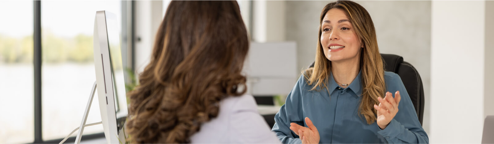 Woman behind a desk talking to another woman with her back towards the camera